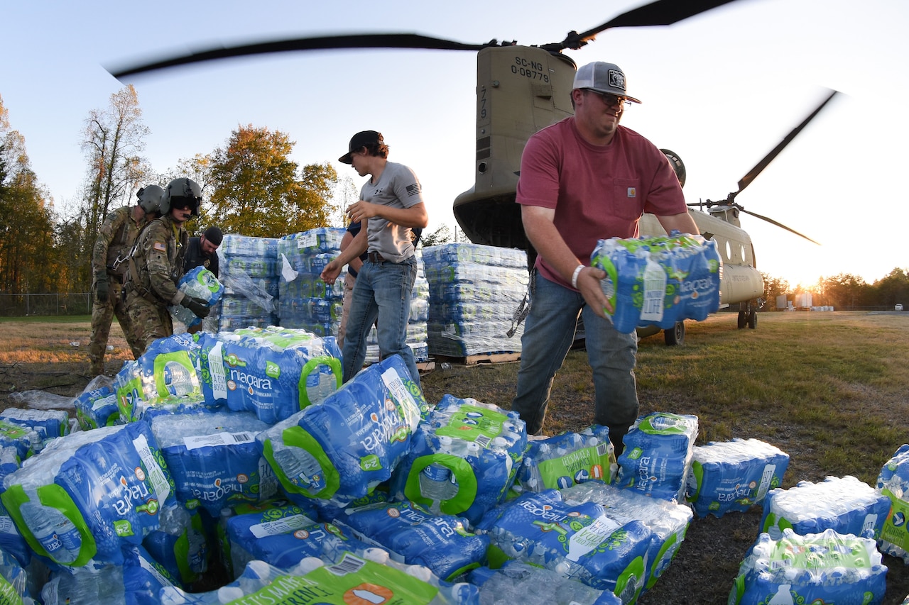 Troops and civilians pass packages of bottled water they are unloading from a parked helicopter in a field.