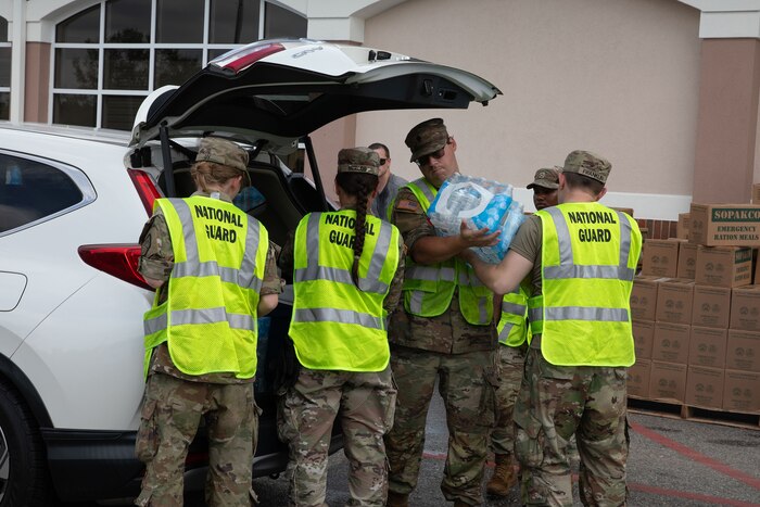 A group of soldiers wearing yellow vests that read "National Guard" load bottled water into the back of a car during daylight.