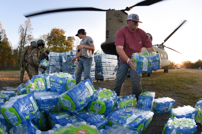 Troops and civilians pass packages of bottled water they are unloading from a parked helicopter in a field.