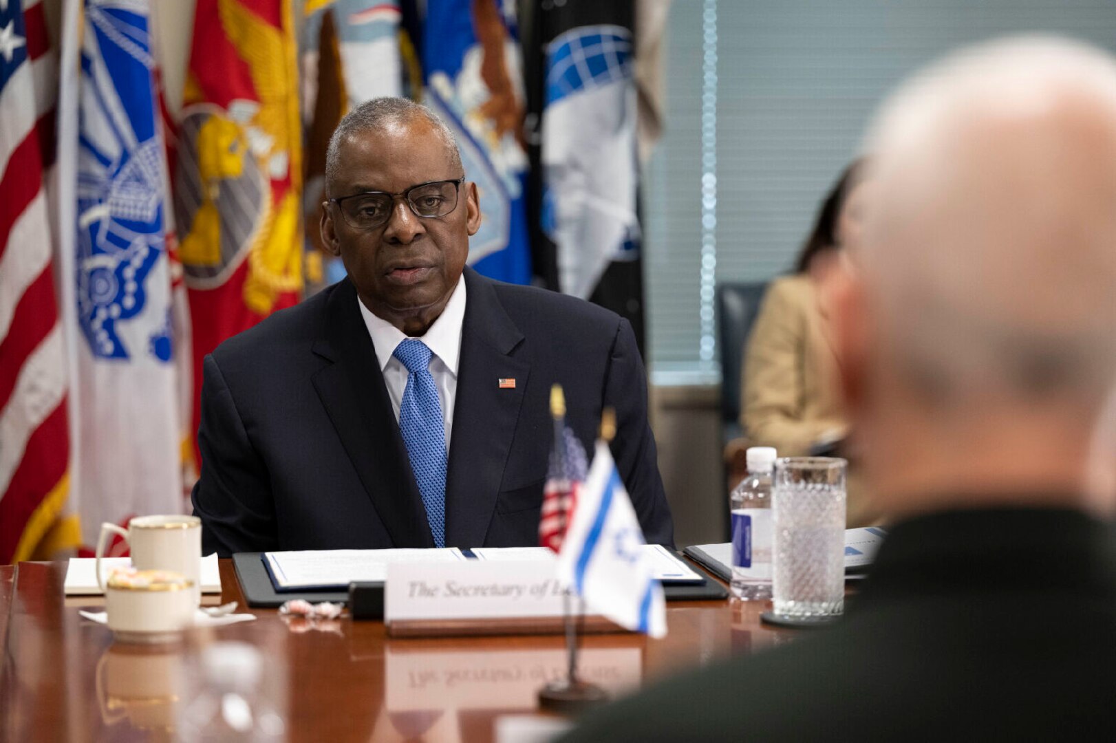 A man in business attire sits at a table displaying U.S. and Israeli flags.