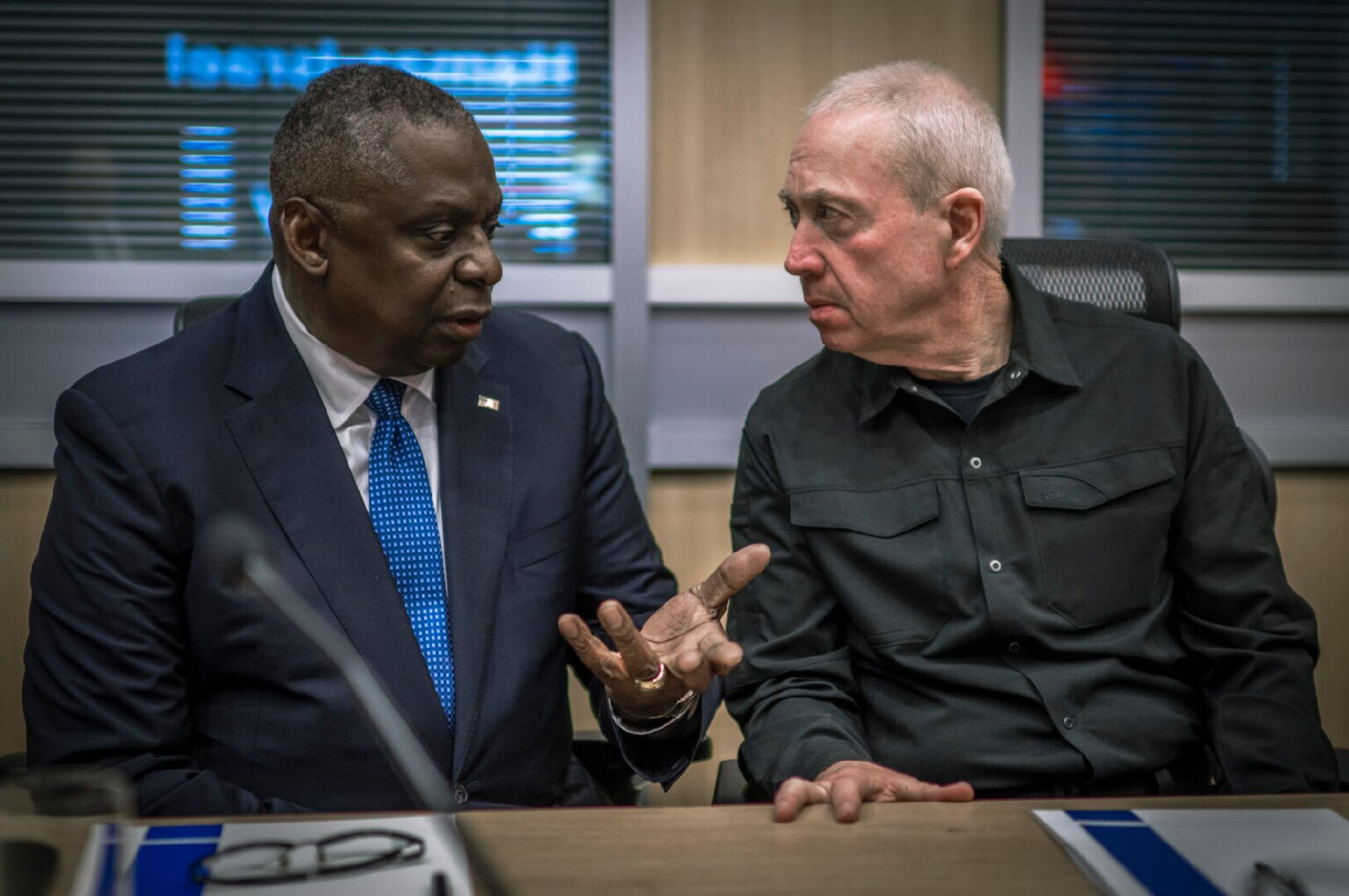 Two men in civilian attire talk while seated at a table.