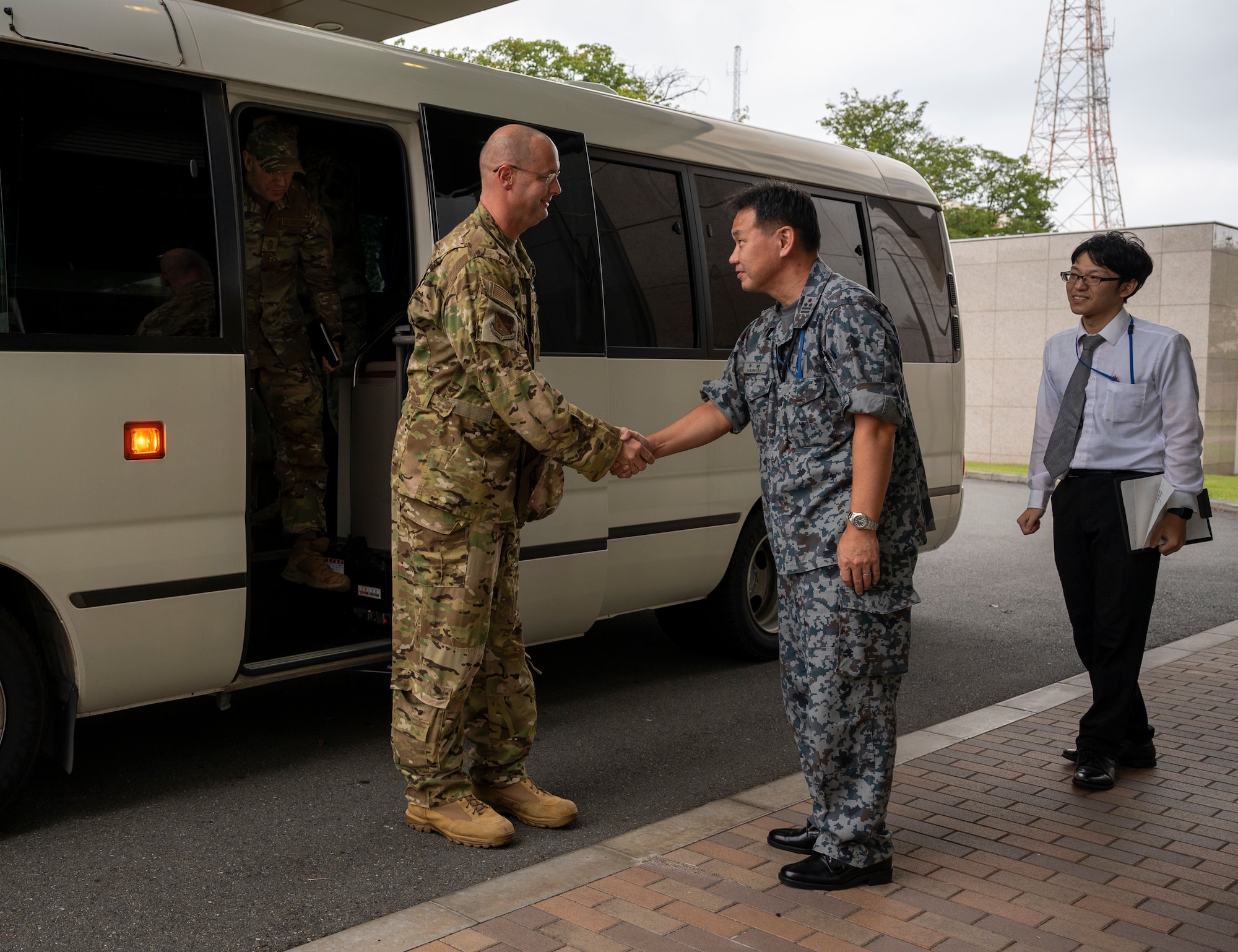 A U.S. and Japanese military member shake hands in front of a white bus.