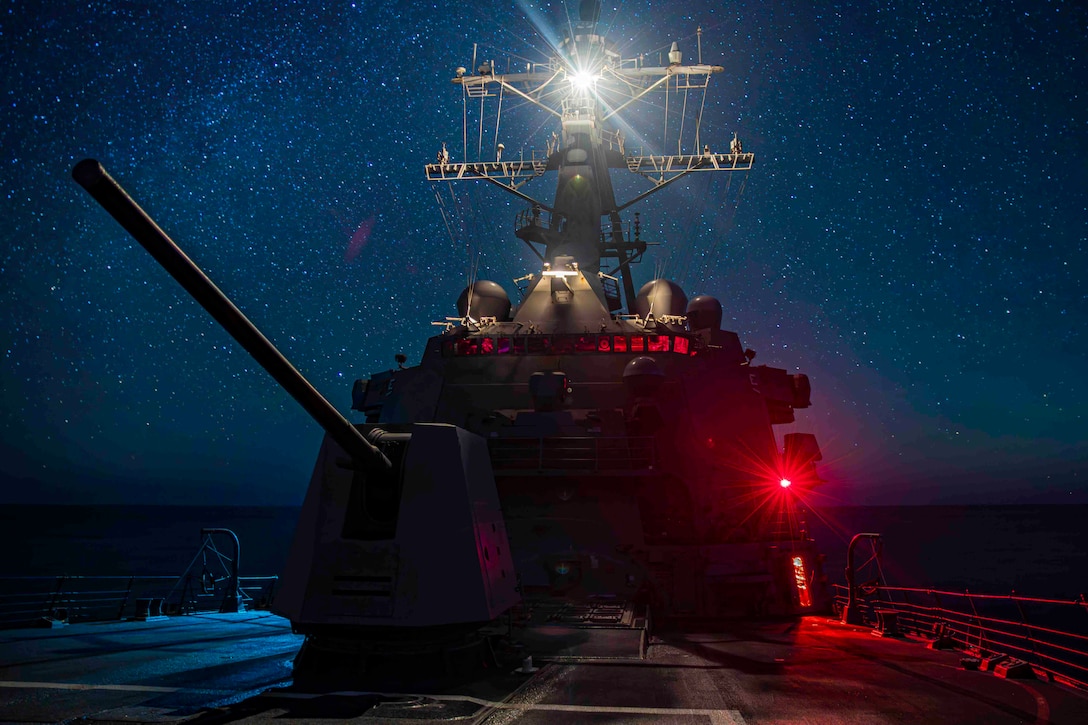 A military ship with its red and white navigation lights on sails at night under a starry sky.