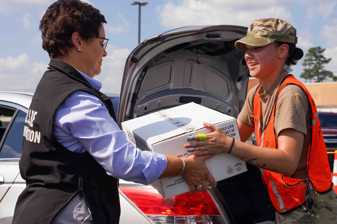 A soldier and a civilian hold a box of food supplies. A car with an open trunk is in the background.