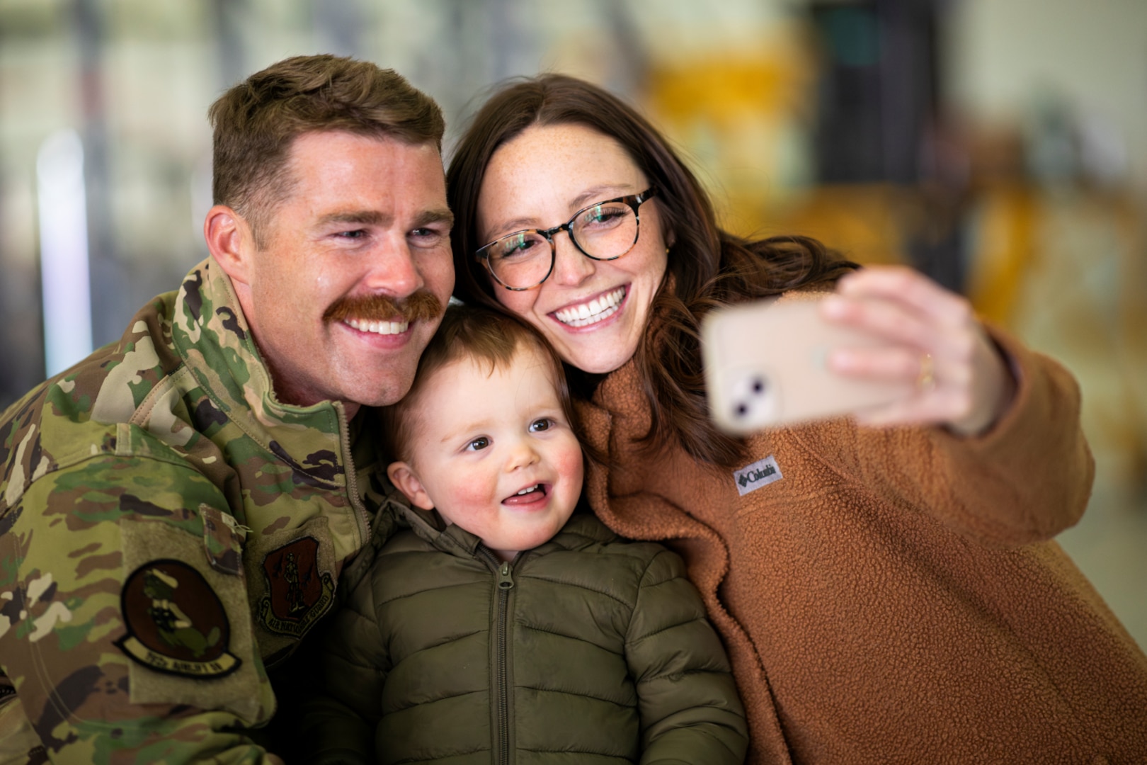 Uniformed service member smiles with his wife and toddler as they pose for a selfie.