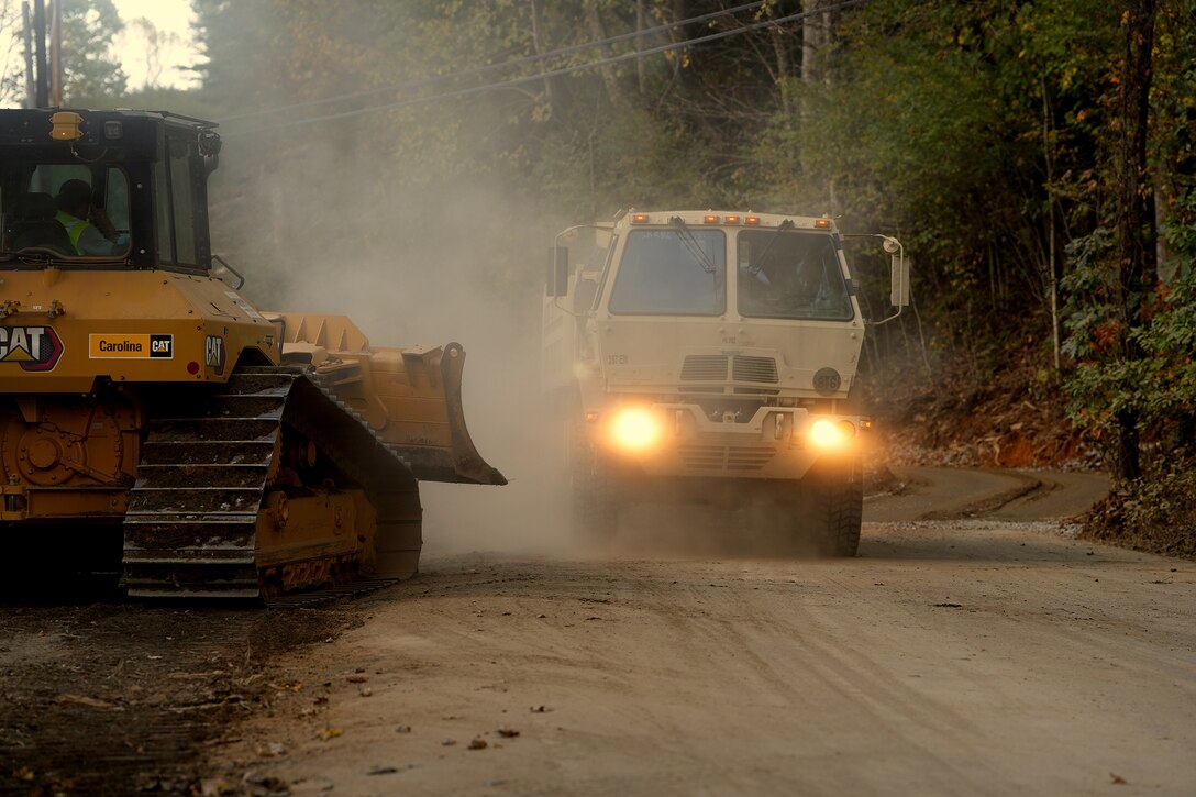 A dump truck and a plow operate on a dusty road on a hazy day. Headlights from the truck illuminate the photo.