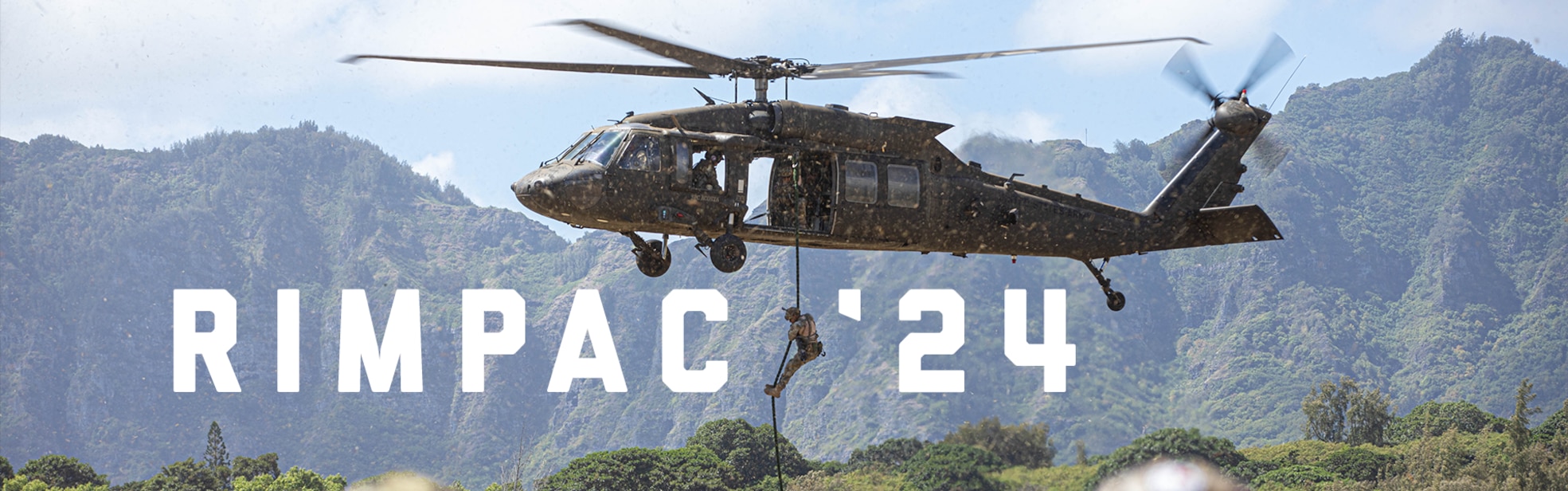 A photo of a person roping down from a helicopter. There are mountains in the background. The words RIMPAC '24 are written onto the photo.