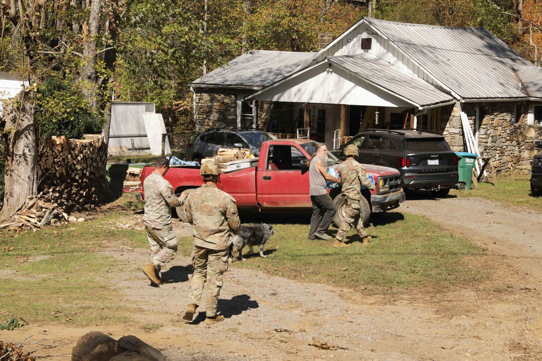 Soldiers walk with a civilian towards a house while carrying cases of water during the daylight.