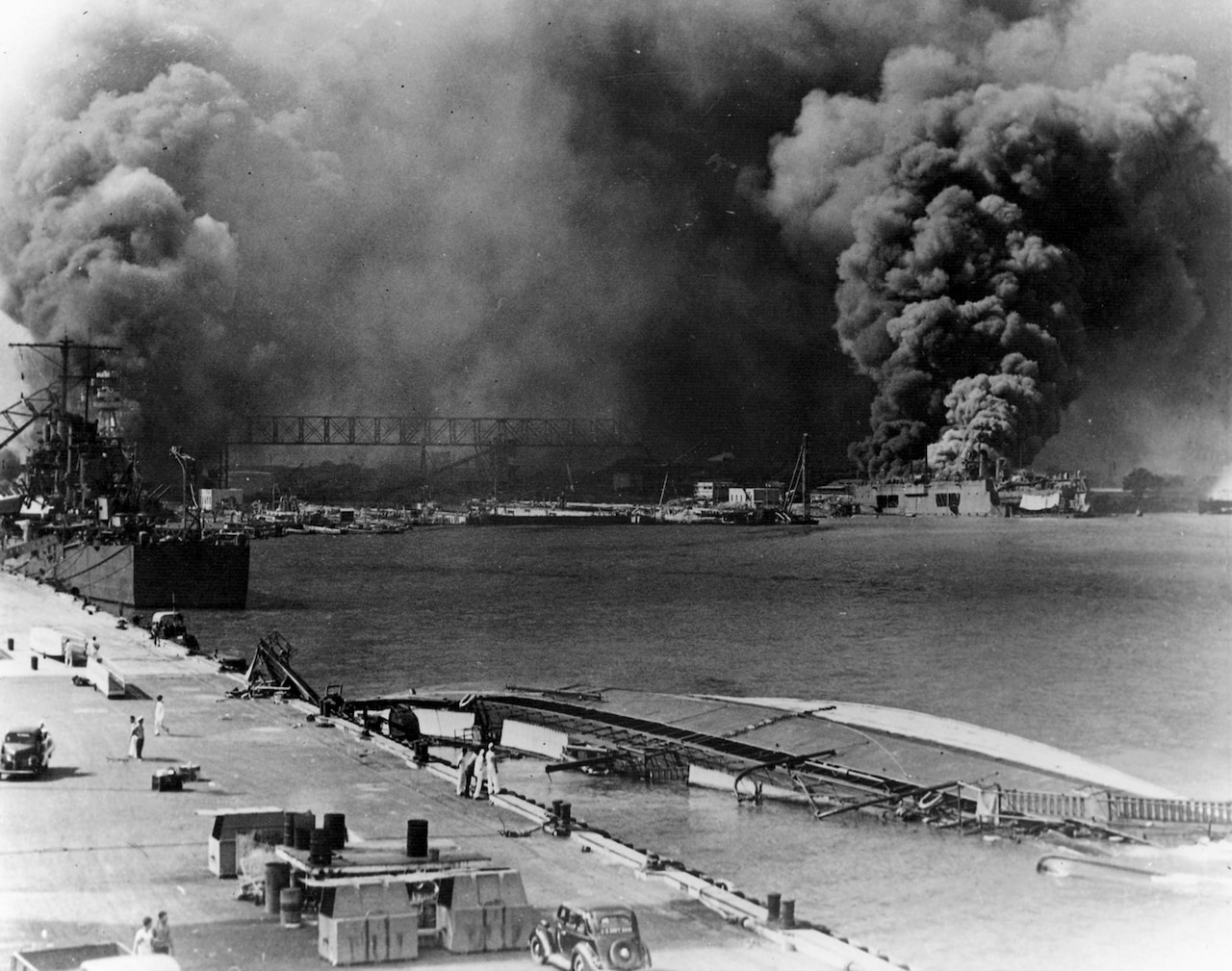Multiple ships explode and sink. Smoke rises over a harbor.