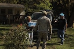 A woman stands outside holding a case of water bottles while a Soldier and man look on.