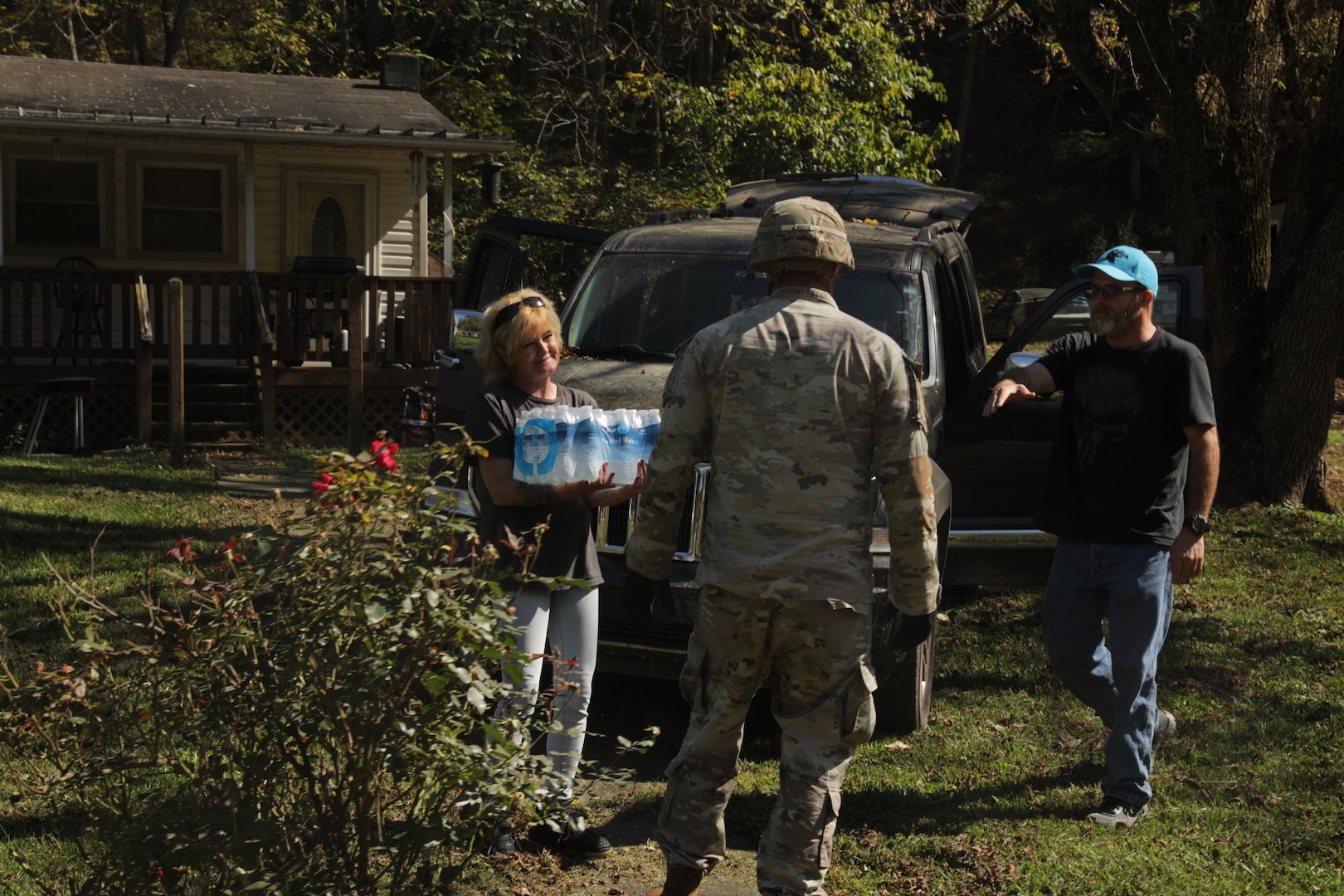 A woman stands outside holding a case of water bottles while a Soldier and man look on.