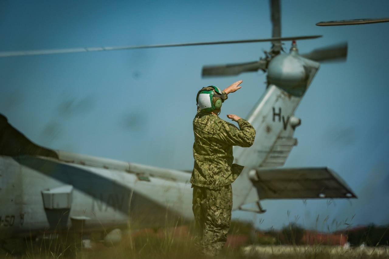 A service member directs a helicopter as it takes off during the day.