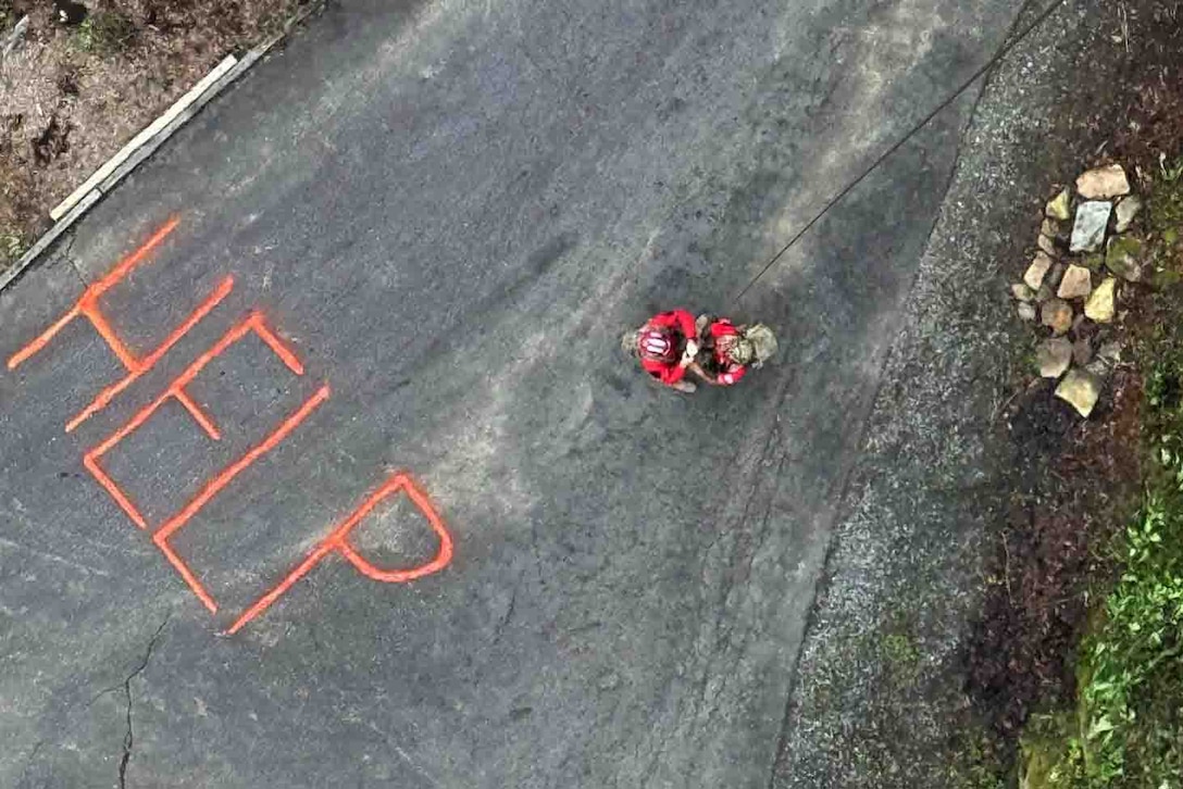 Two airmen rappel down from a helicopter to a driveway with the word ‘HELP’ written on it in large red capital letters.