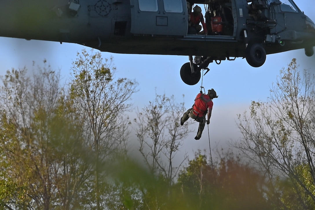 An airman holds a cable while being lowered from a helicopter in a wooded area.