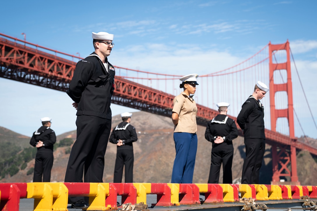 Sailors and Marines stand in formation on the flight deck of a ship. A large bridge and bare hills are in the background.