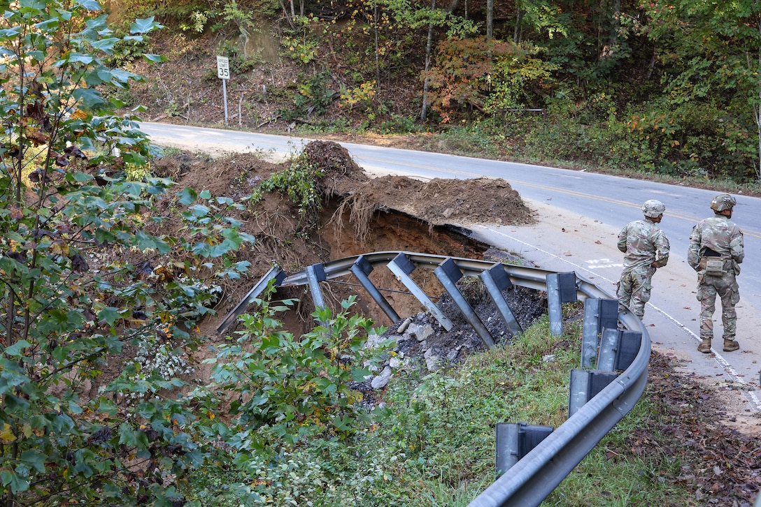 Soldiers assess hurricane damage to a curved road and guardrail during daylight.