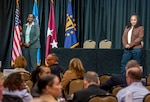 U.S. Army Lt. Gen. Telita Crosland and U.S. Air Force Chief Master Sgt. Tanya Johnson stand on a stage in front of a table and address audience.