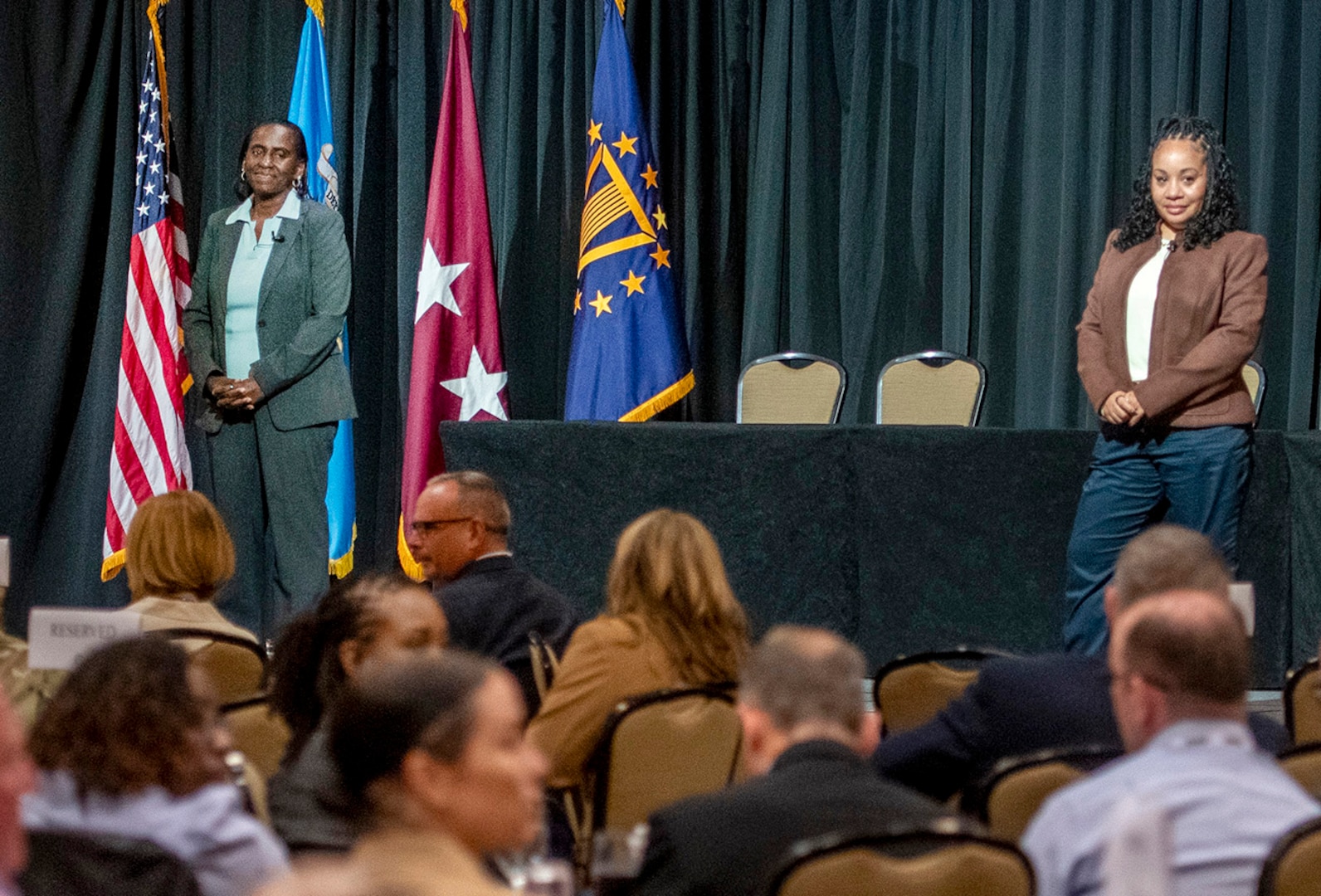 U.S. Army Lt. Gen. Telita Crosland and U.S. Air Force Chief Master Sgt. Tanya Johnson stand on a stage in front of a table and address audience.