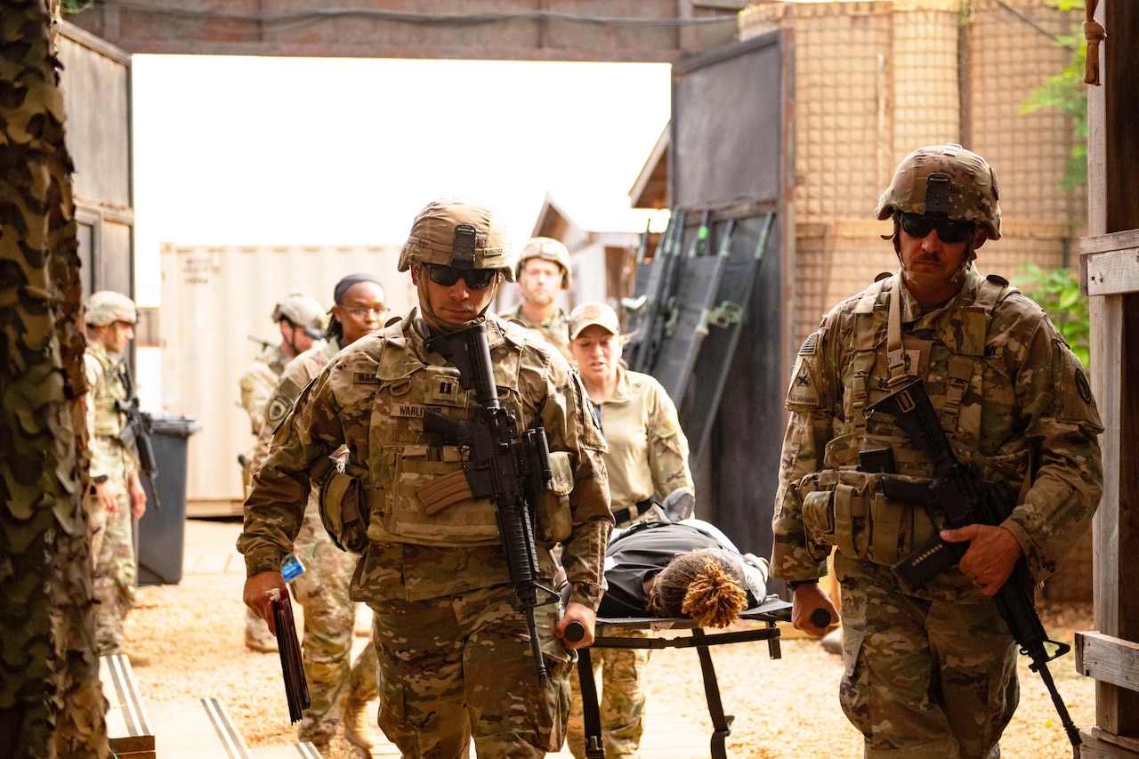 Four soldiers carry a victim on a stretcher while several others look on.