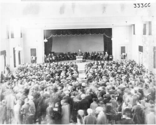 Black and White photo of a ceremony held in an auditorium with a group of men and women standing. Someone is on a stage standing behind a podium with people sitting behind them.