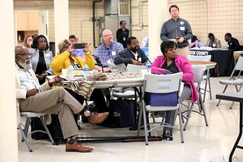 Group of community members listen to presentations from other outreach groups and government agencies on procedures and available resources to them with some taking photos.