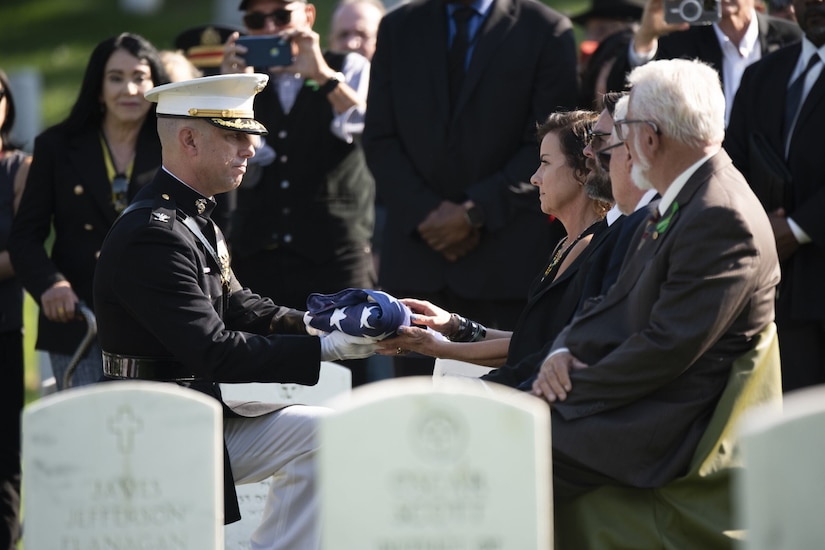 A man kneels in front of a seated woman to hand her a folded flag.