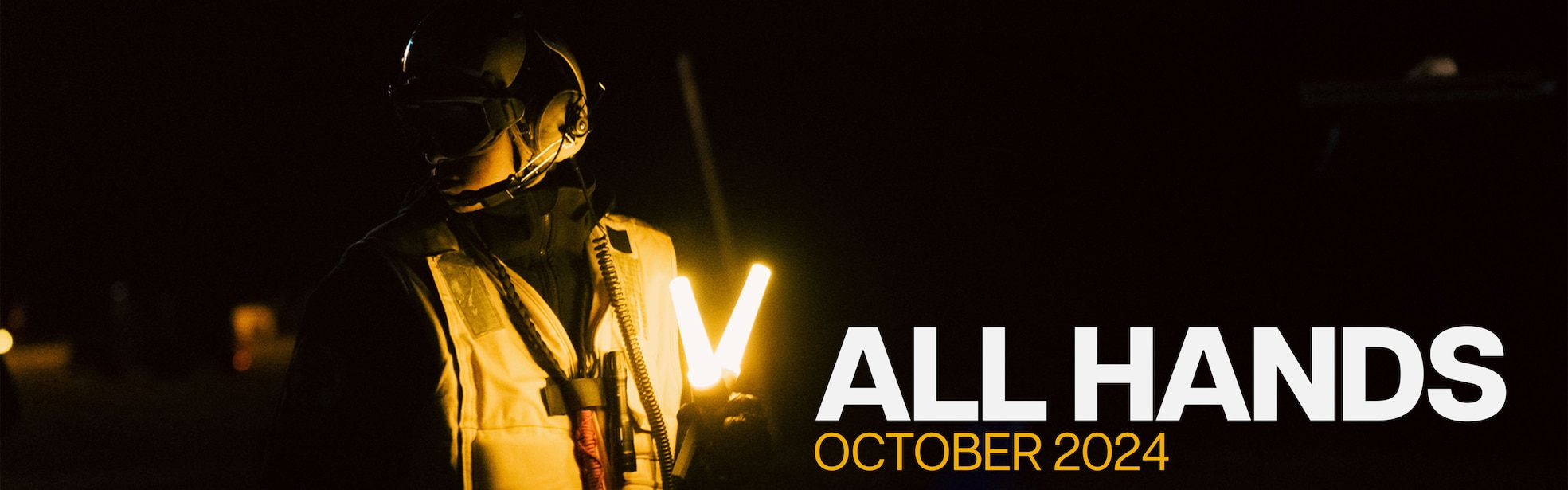 Sailor in their airman vest holding glowing yellow sticks at nighttime on the ship. White font titled All Hands with orange font stating October 2024