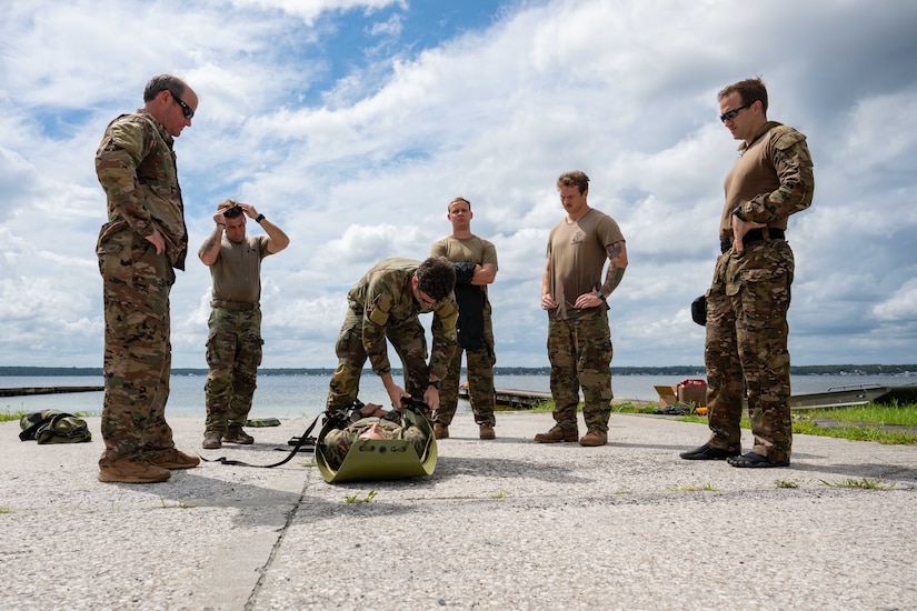 A group of soldiers observe training as another soldier secures a volunteer onto a stretcher on the ground.