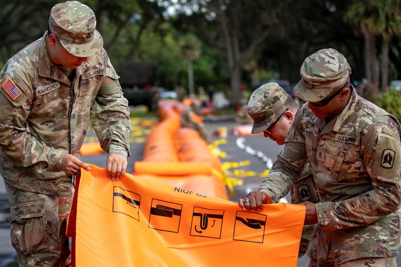 Three service members deploy an orange flood mitigation device that rests on top of asphalt.