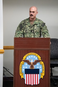 A Navy officer in camouflage speaks behind a podium with the Defense Logistics Agency logo on it