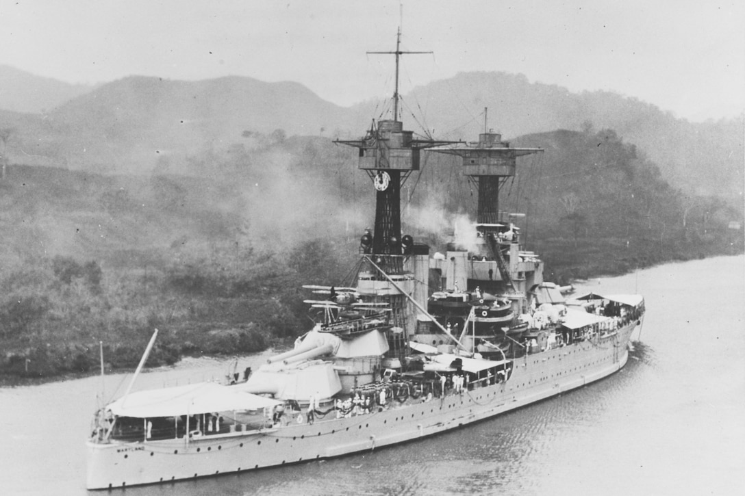 Black and white photo of a battleship in waters near a shoreline, with mountains in the background.
