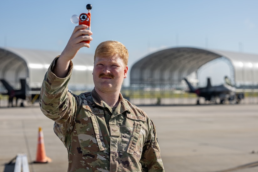 An airman holds up a small red device while standing on a flight line.