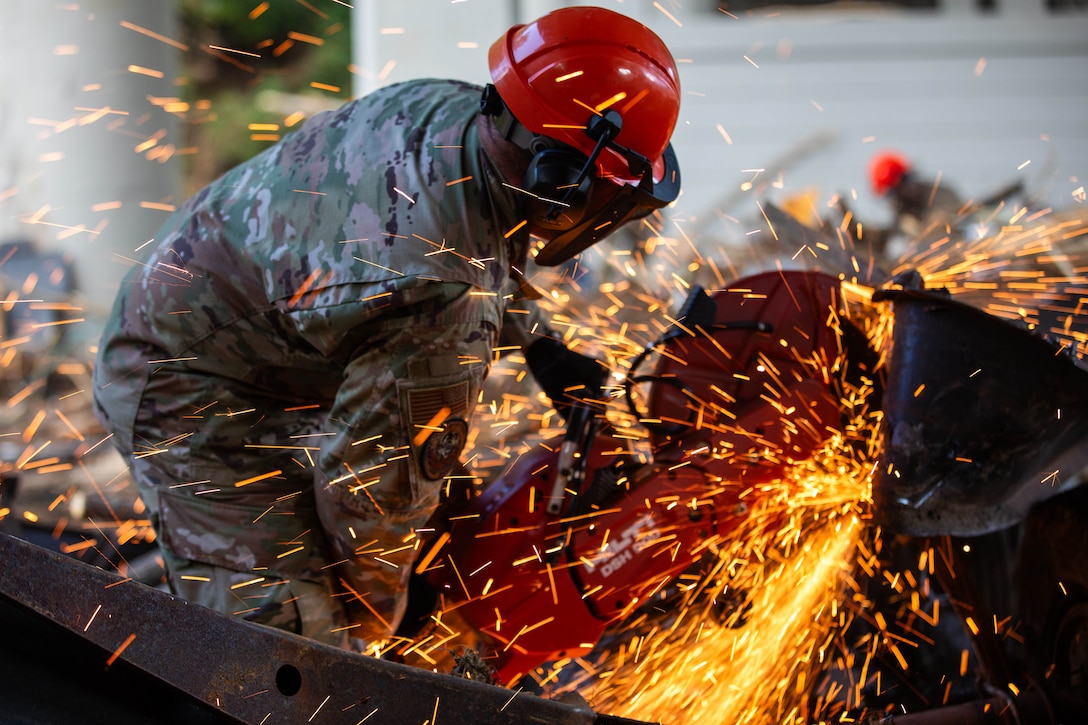 An airman wearing a hardhat and a face shield uses a chainsaw to cut through metal debris creating lots of orange sparks.