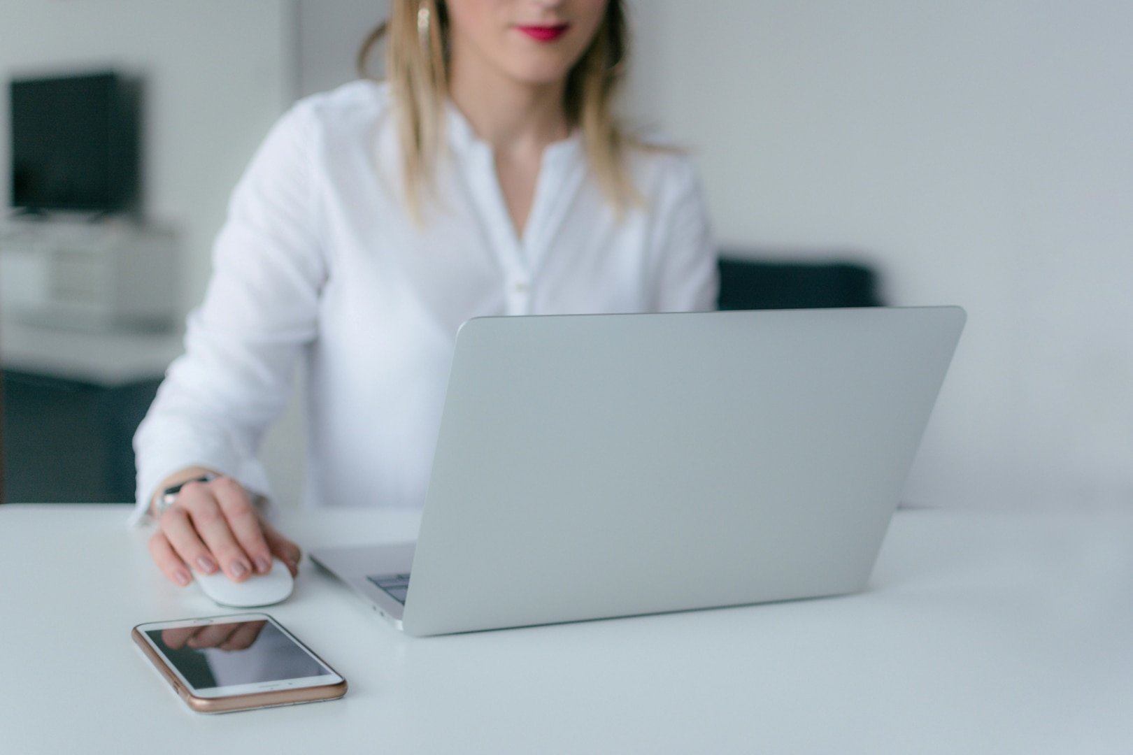 Woman sits at table looking at laptop with smartphone next to her.