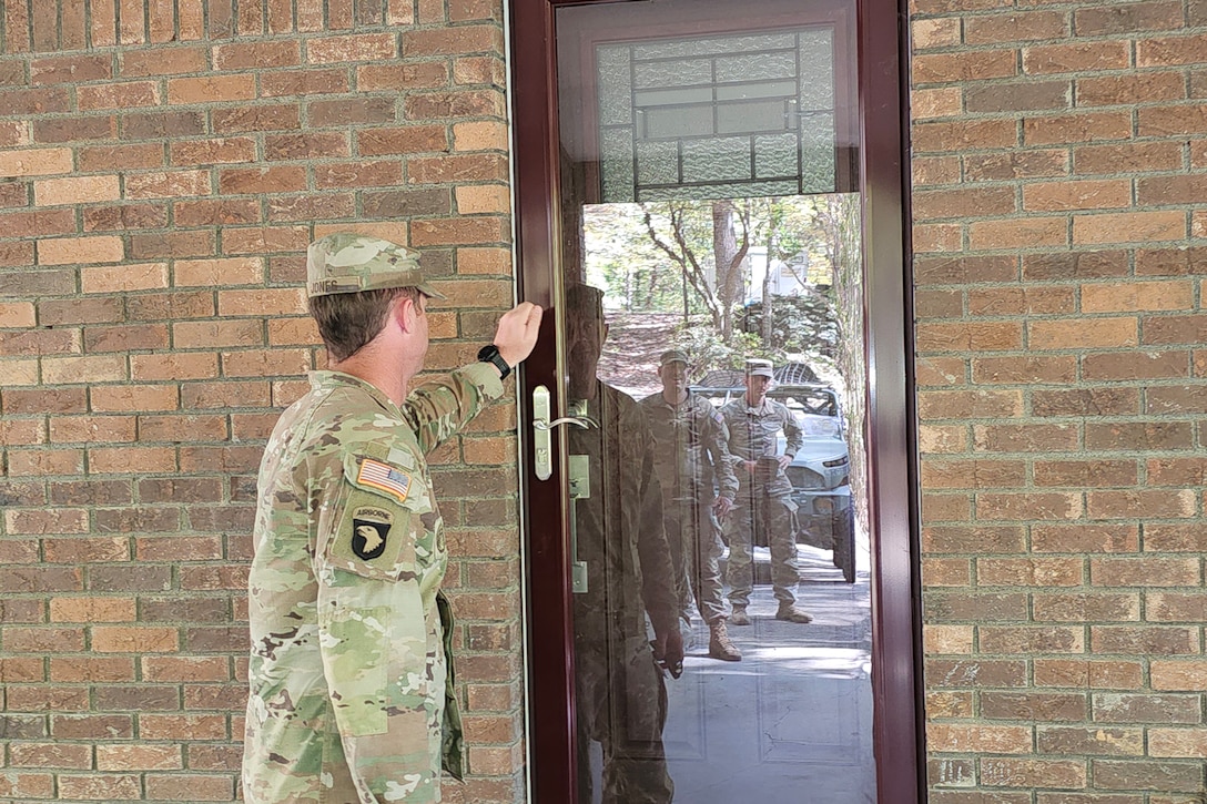 A soldier knocks on a glass storm door during daylight as the reflections of two other soldiers can be seen in the door.