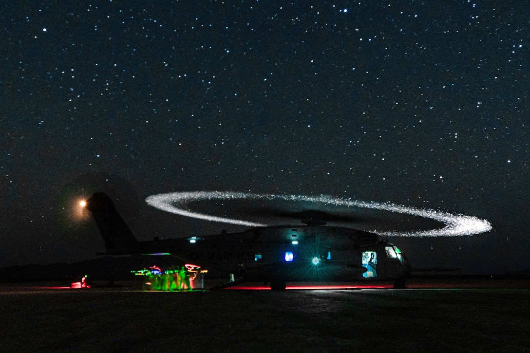 Marines unload cargo from a helicopter on a starry night as the helicopter emits white, red and green lights. The moving blades of the helicopter create a white halo above it.