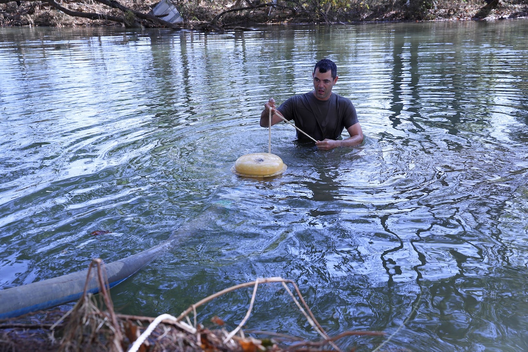 A sailor stands in waist-high water while holding a water purification device on a rope during daylight.