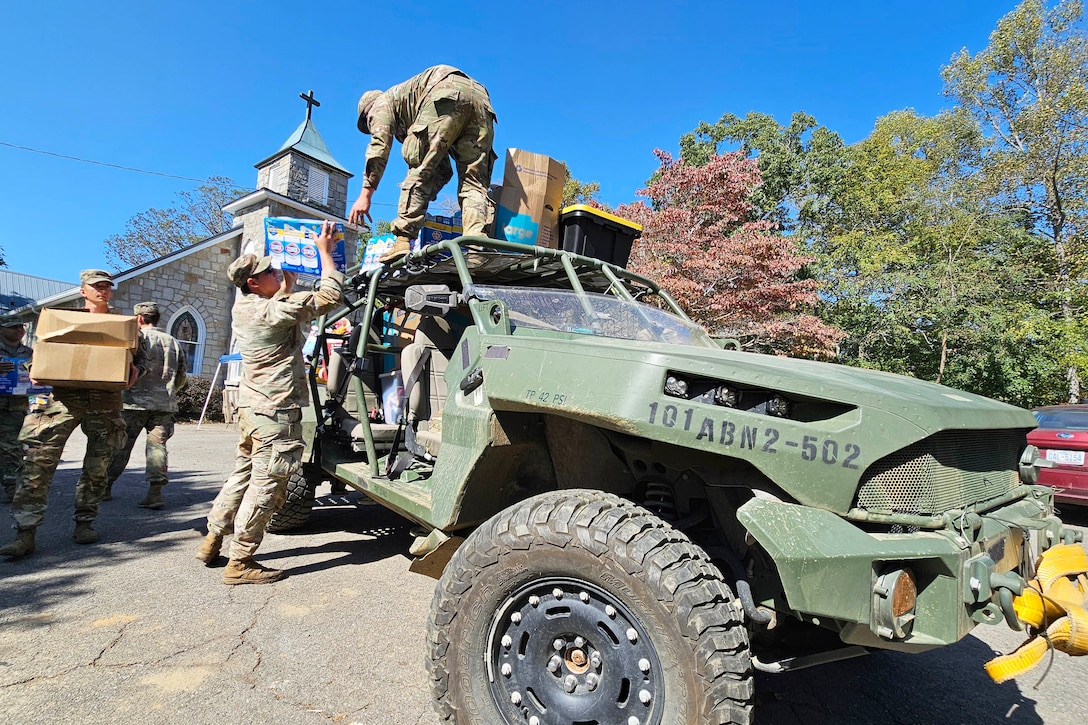 A soldier hands a box to a soldier standing on top of parked vehicle in front of a church as fellow soldiers line up with boxes.