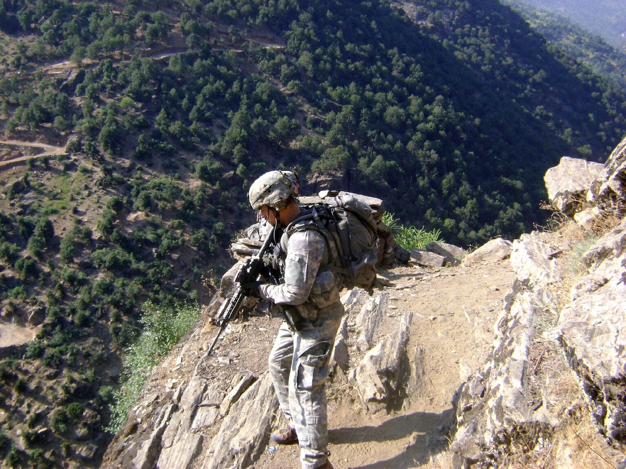 A man in full combat uniform and rifle stands on the side of a mountain looking at the valley below.