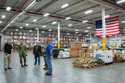 Five men stand talking in a warehouse with an American flag hanging in the background. There are pallets of boxes on the ground and shelving stocked with boxes in the background.