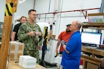 Two men stand talking inside of a warehouse with a table and packing boxes in front of them. One of the men is wearing a camouflage military uniform. There are people in the background.
