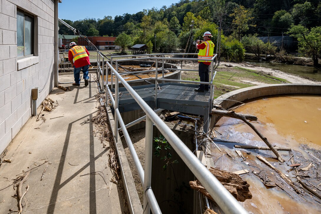 Two men stand on a walkway above wastewater holding tanks filled with brown water and woody debris.