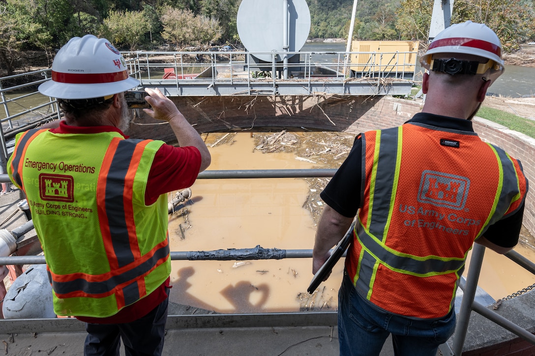 Two men wearing hard hats and safety vests stand above a wastewater treatment container that is full of brown water.