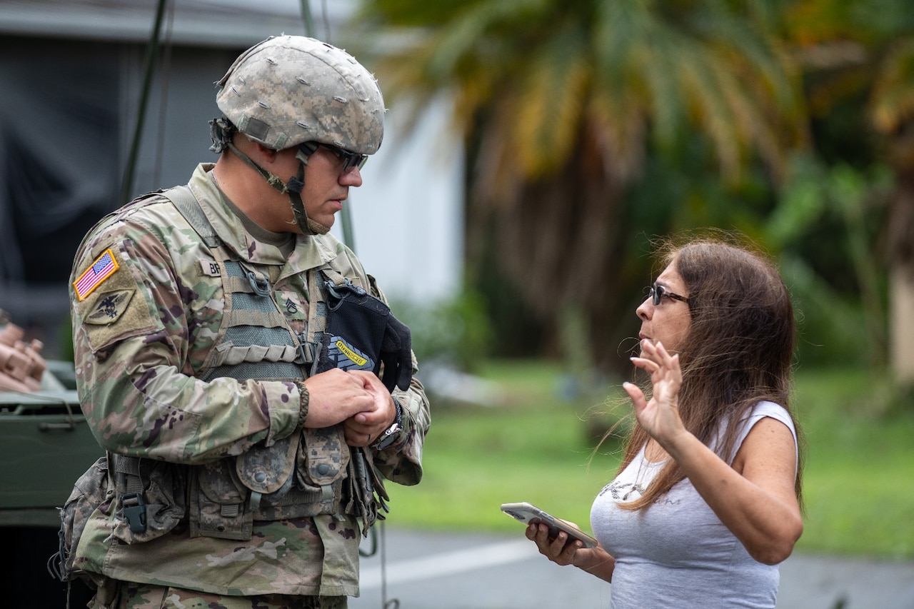A Guardsman talks with a resident who is holding a phone; palm trees and a road are in the background.