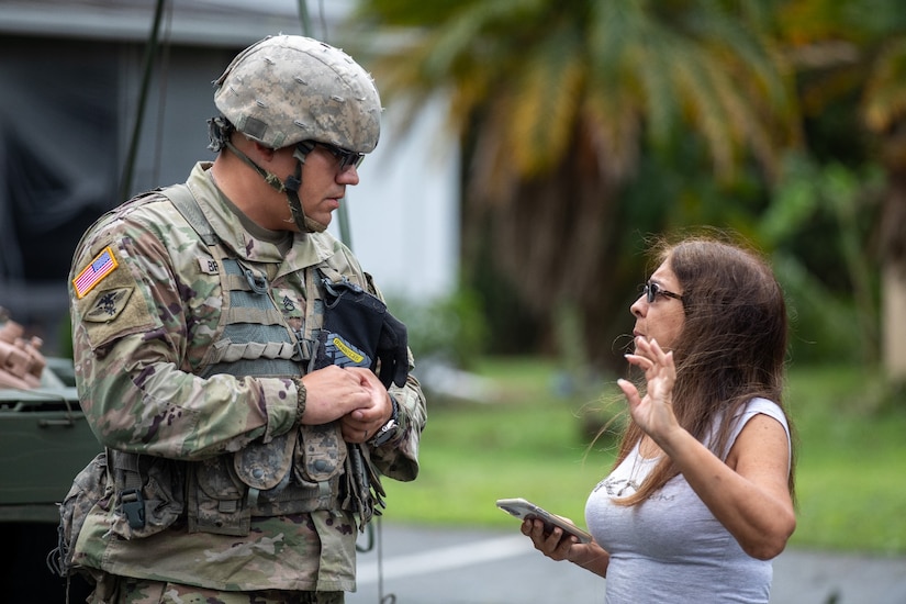 A Guardsman talks with a resident who is holding a phone; palm trees and a road are in the background.