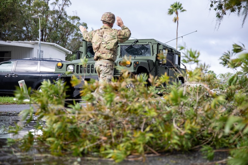 A guardsman standing by the road signals the driver of a green military vehicle as it drives by a house with a van parked in front.