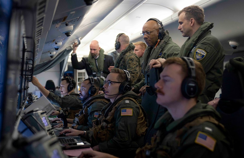 A service member in flight gear observes service members as they work on computer stations aboard a plane.