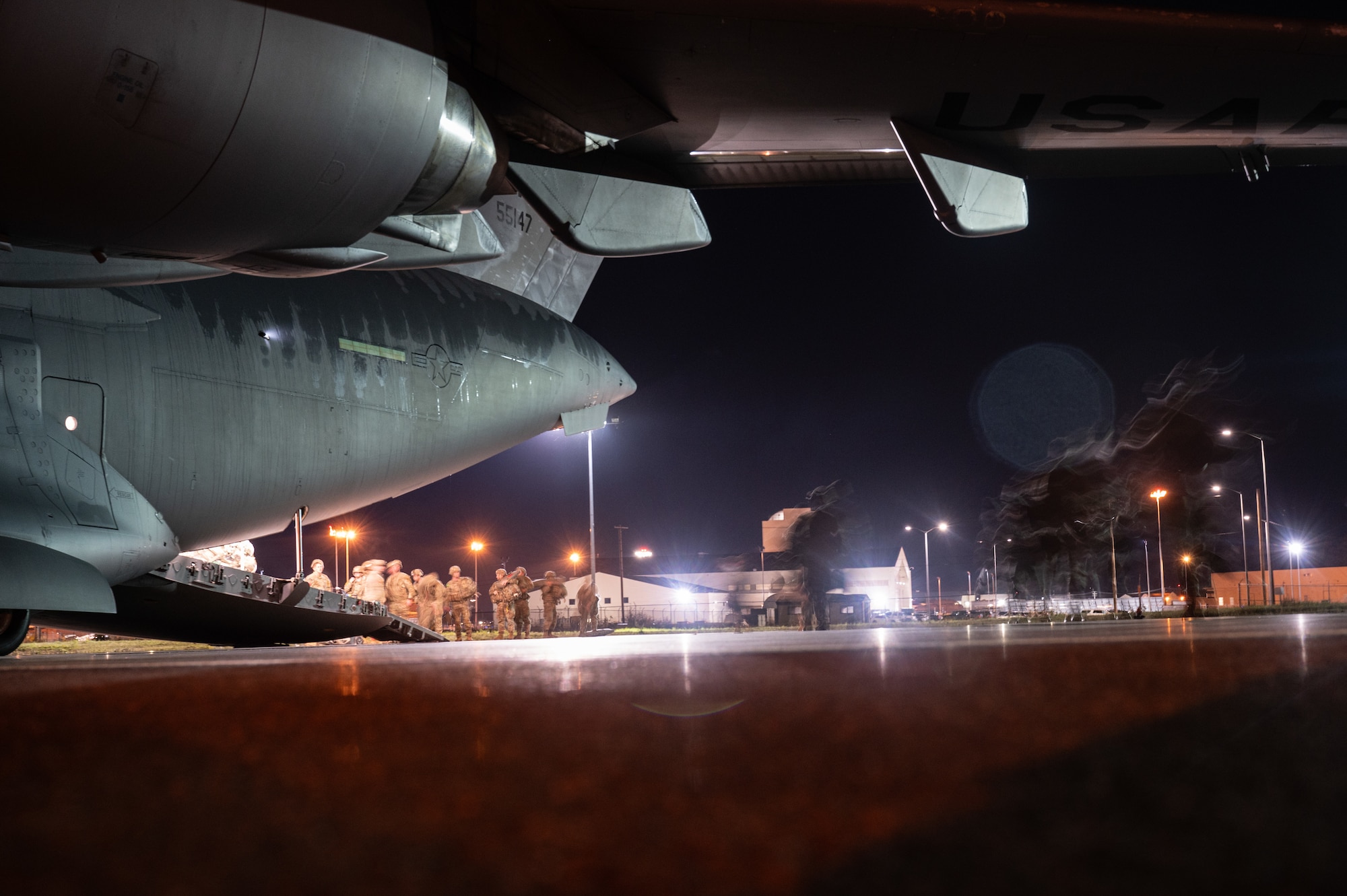 U.S. Army Soldiers walk onto a C-17 Globemaster III aircraft from the back ramp while wearing parachutes at night.