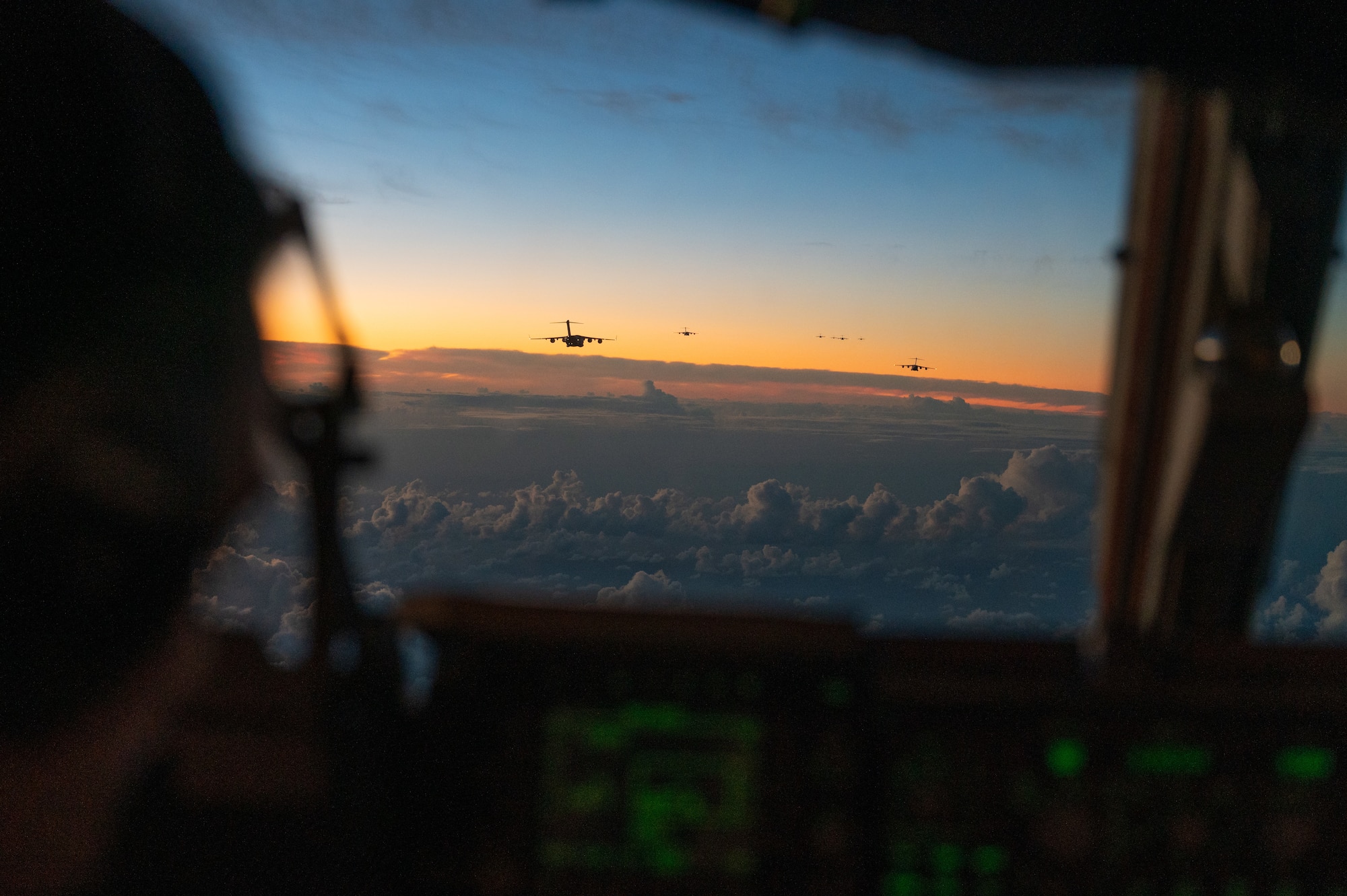 Pilot looks through a window and sees a C-17 Globemaster III aircraft flying off in the distance during sunrise.