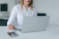 Woman sits at table looking at laptop with smartphone next to her.